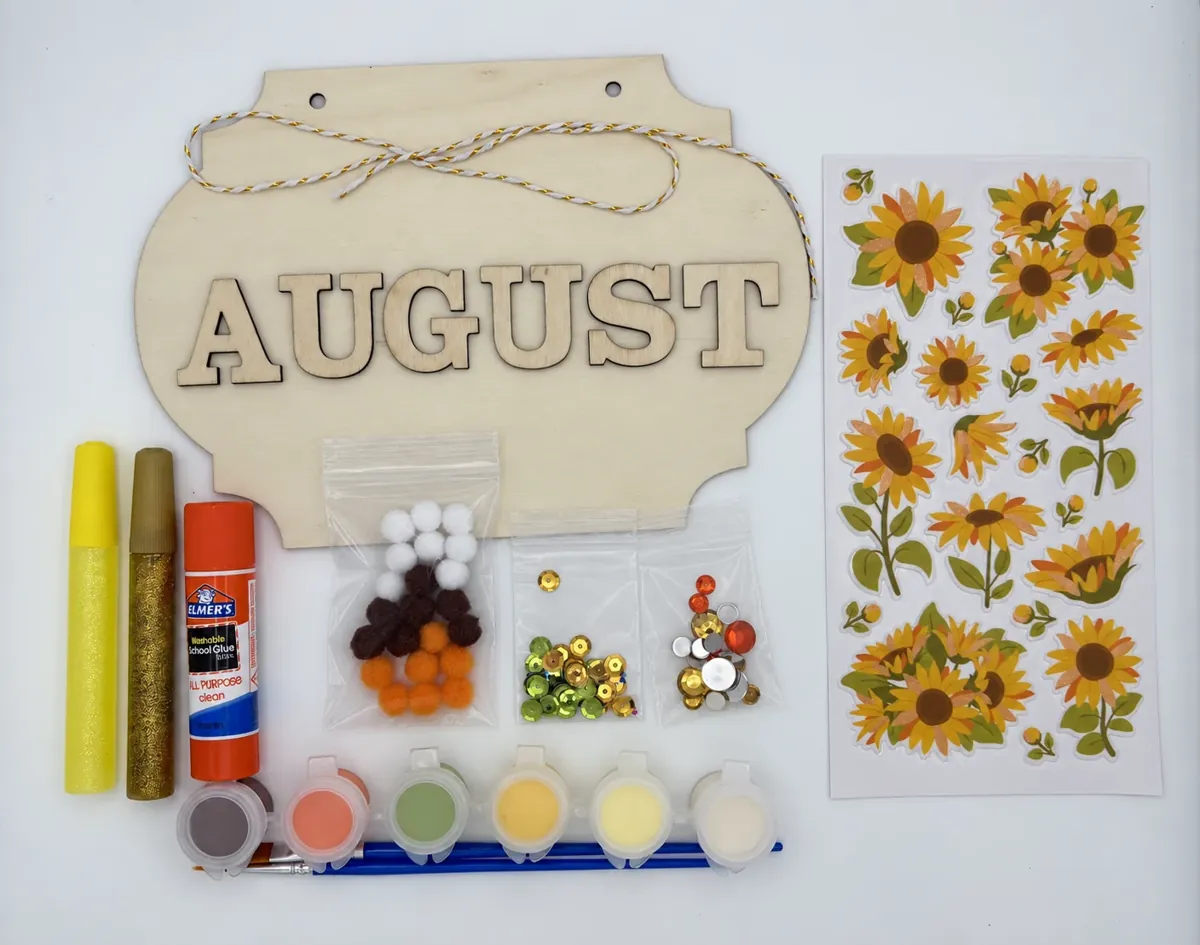 Sunflower kit contents laid out with the AUGUST plaque, paints, brushes, glue, gems, pom-poms, and stickers
